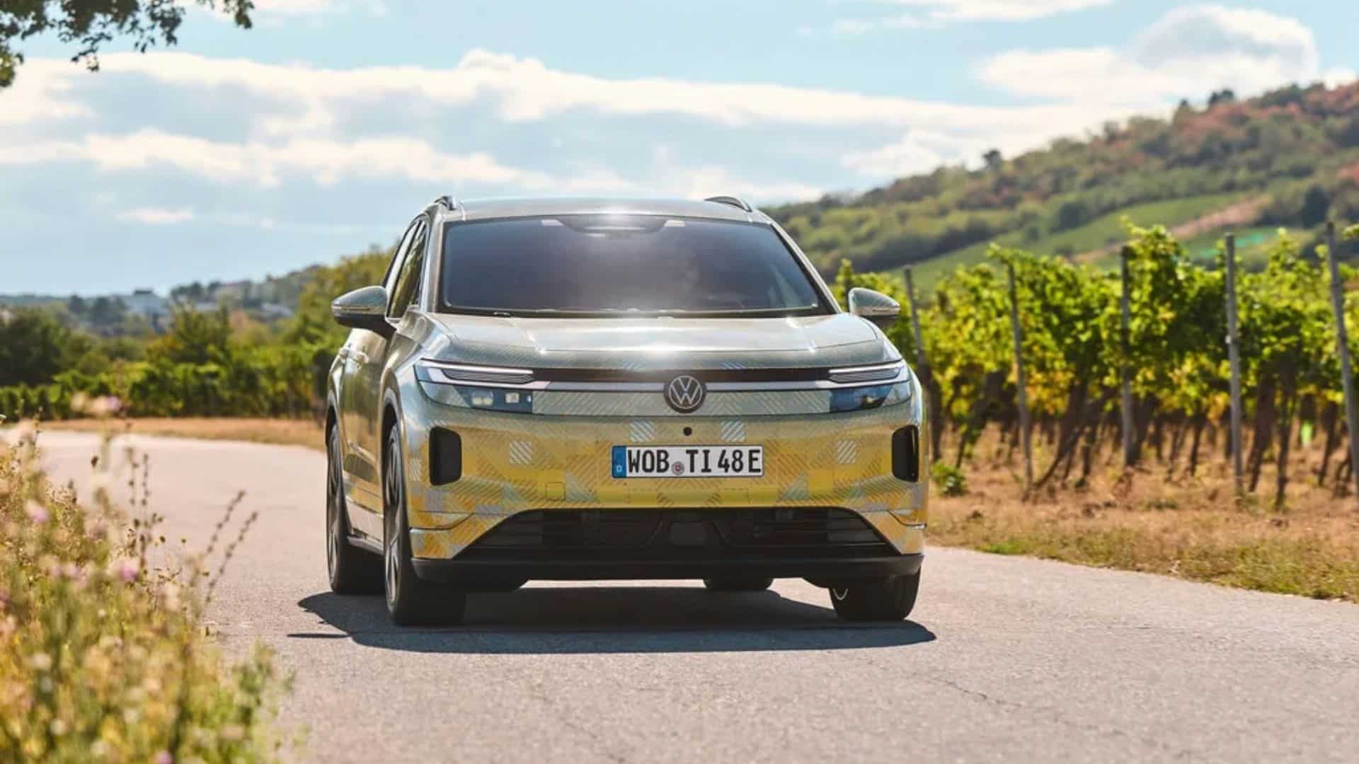 Protótipo camuflado do Volkswagen ID. Tiguan 2027 dirigindo em uma estrada rural ladeada por vinhedos. A dianteira exibe faróis de LED conectados por uma faixa iluminada, o logotipo da VW ao centro e para-choque com detalhes em pre