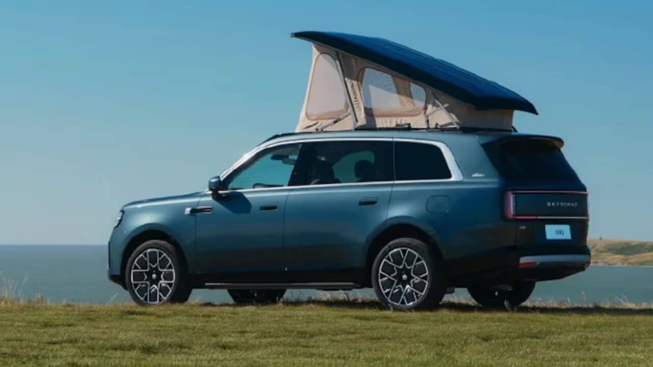 SUV Xiaomi SkyNomad N90 Max Studio na cor azul-petróleo, visto de perfil, estacionado em um campo gramado à beira-mar. O veículo possui uma barraca de teto cinza e azul armada sobre ele. O céu está limpo e azul