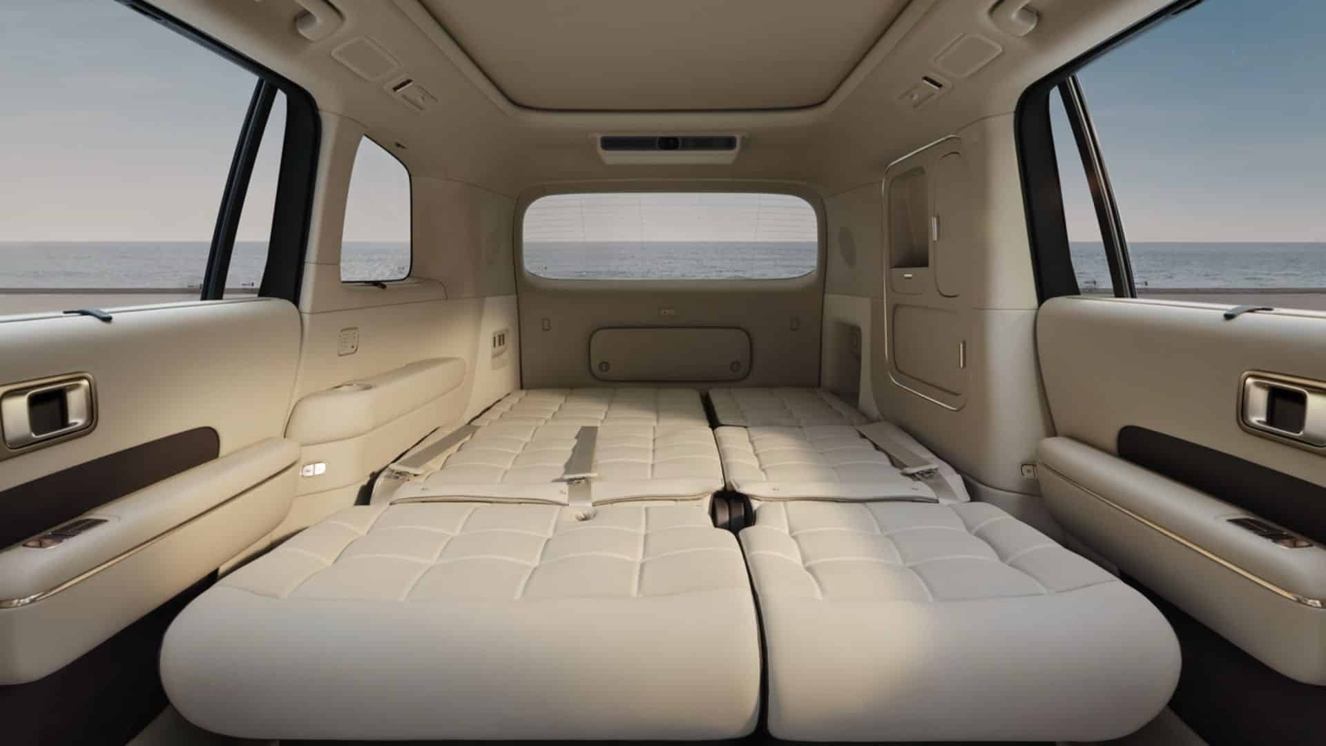 Vista interna do porta-malas do Xiaomi SkyNomad N90 Max Studio, mostrando os bancos traseiros totalmente rebatidos, formando uma cama plana e espaçosa com estofamento bege acolchoado. Através das janelas, é possível ver o mar e o horizonte