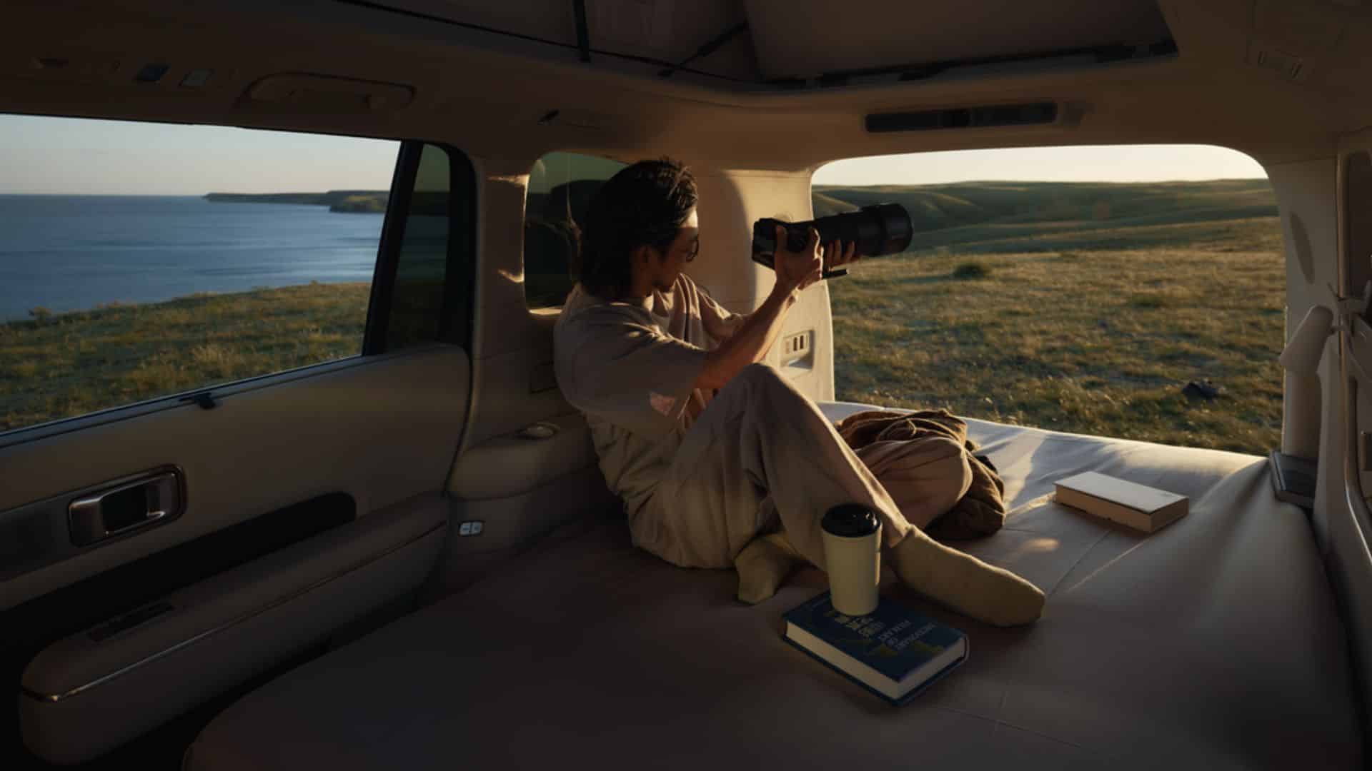 Um homem sentado na parte traseira do Xiaomi SkyNomad N90 Max Studio, que foi convertida em área de descanso. Ele está usando roupas confortáveis e segurando uma câmera com lente teleobjetiva, observando a paisagem costeira pela janela durante o pôr do sol. Há um copo térmico e um livro sobre a superfície de dormir