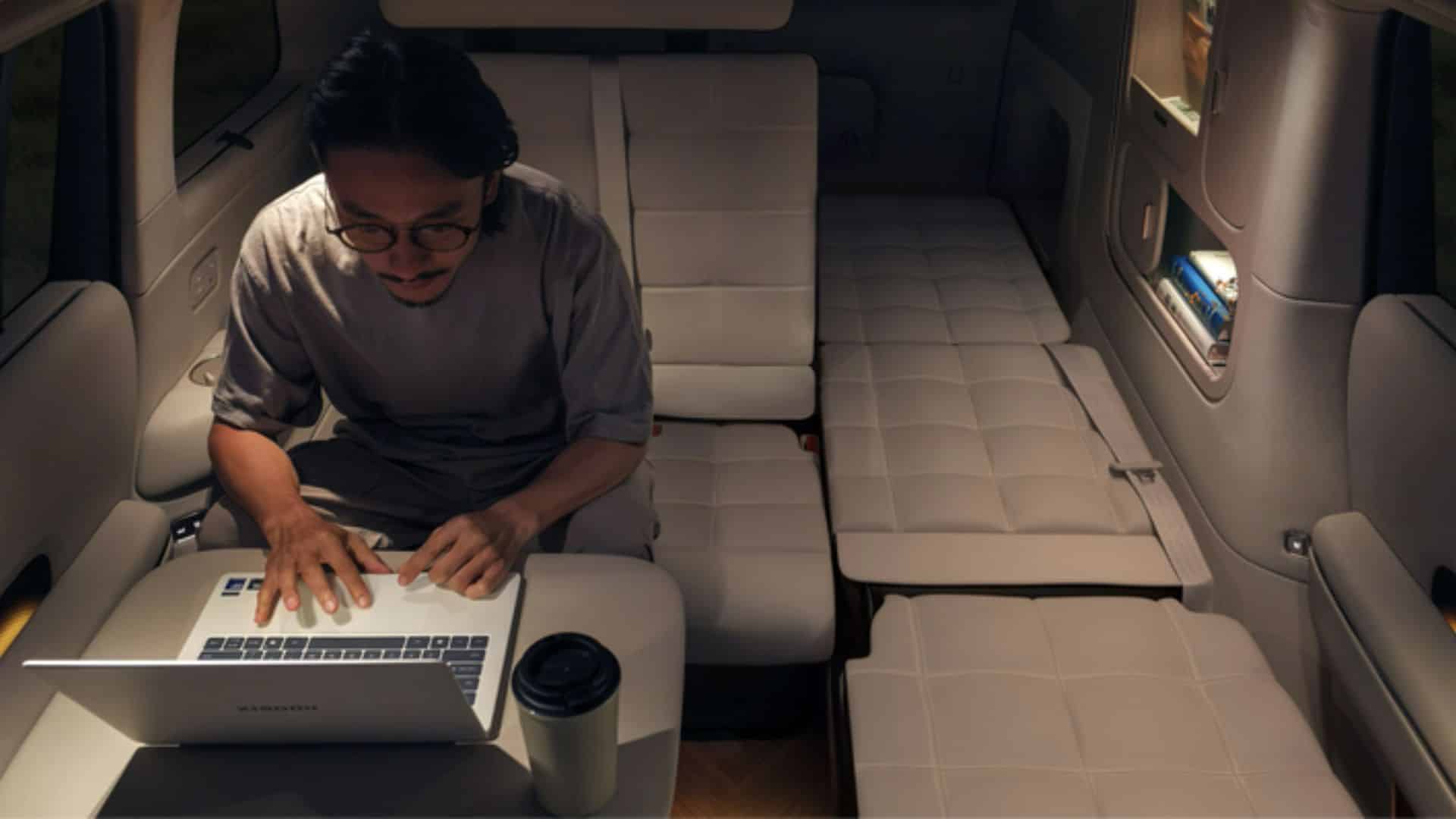 Vista noturna do interior do Xiaomi SkyNomad N90 Max Studio. Um homem de óculos trabalha em um laptop apoiado sobre uma mesa dobrável, enquanto os bancos traseiros estão configurados como camas individuais. O ambiente é iluminado por luzes internas suaves, e há um copo térmico ao lado do laptop