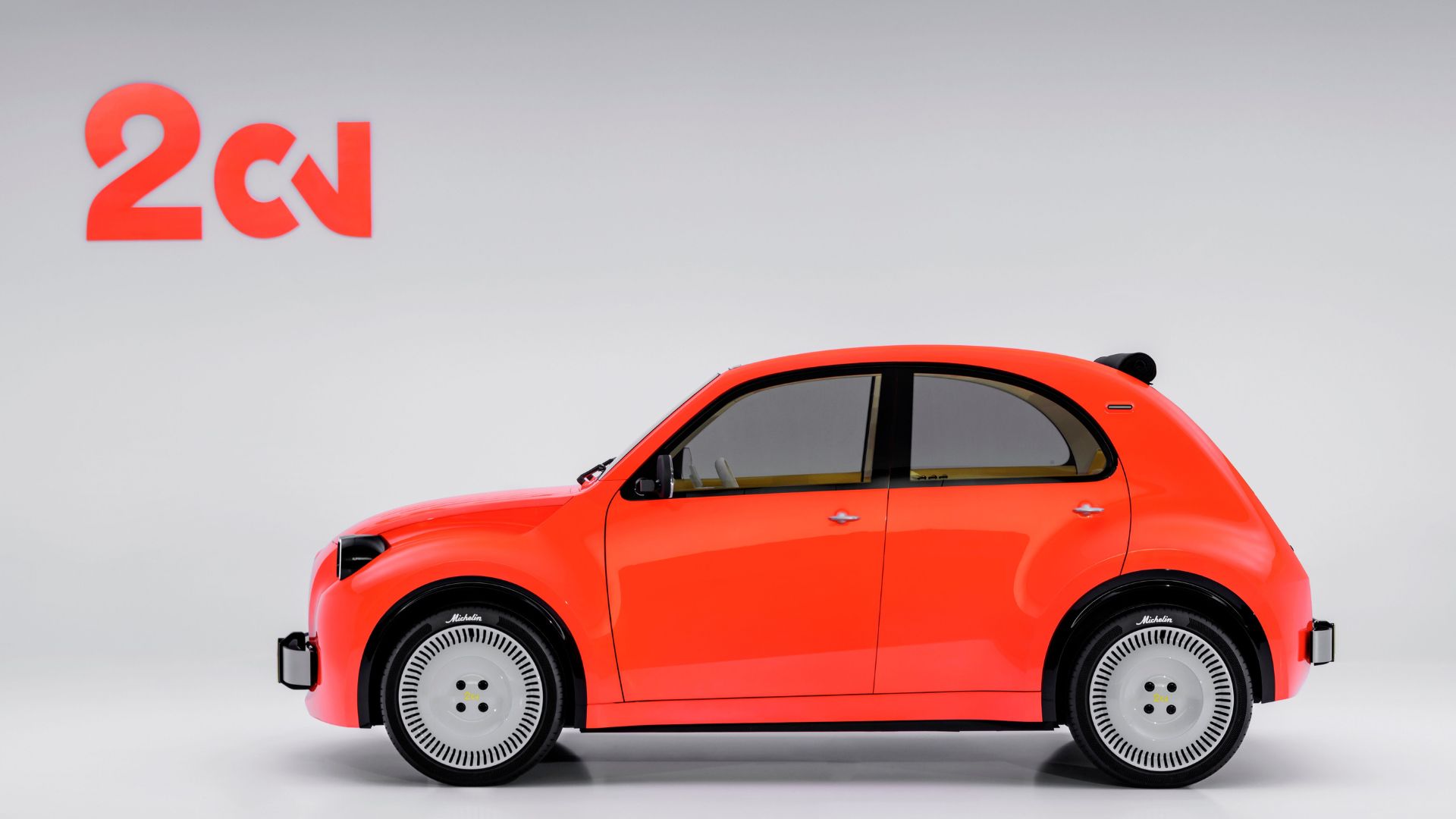 Vista lateral do Citroën 2CV Concept elétrico laranja. O perfil mostra a silhueta compacta e arredondada, com teto de tecido enrolado, maçanetas prateadas e as rodas brancas com detalhes em preto. O logotipo "2CV" aparece no canto superior esquerdo