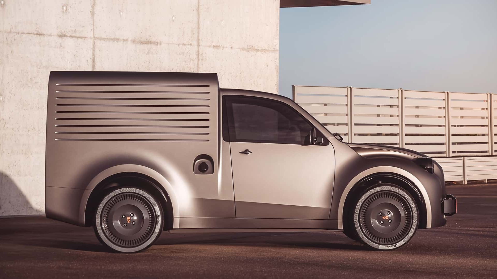 Vista lateral da Citroën 2CV Fourgonette Concept prateada. A imagem destaca a traseira fechada do tipo furgão, com painéis laterais frisados, uma única porta na cabine e rodas escuras com detalhes em vermelho. O fundo mostra uma parede de concreto e uma cerca de madeira