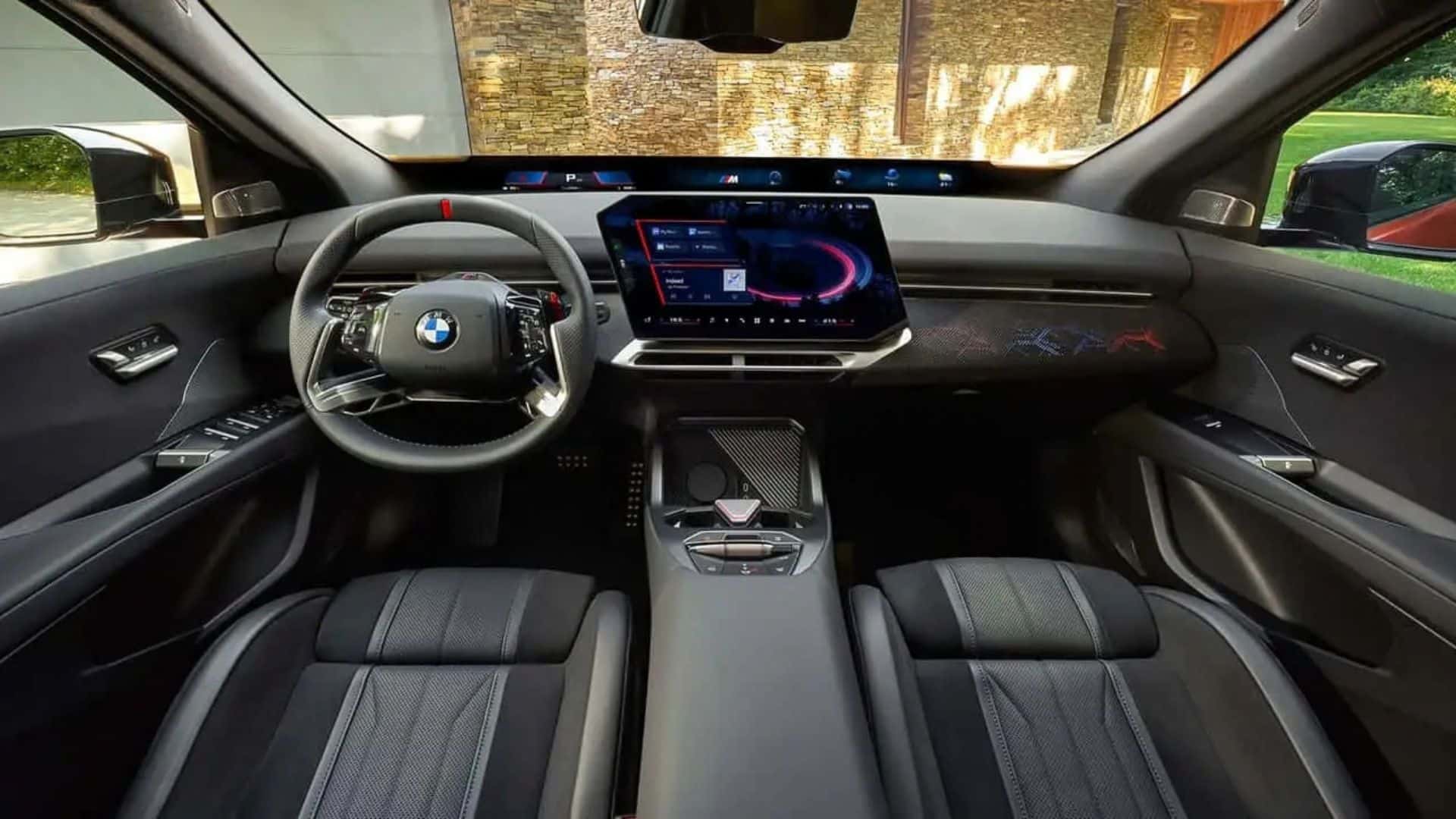 Vista interna do painel e console central do BMW iX4, exibindo o volante esportivo com logotipo BMW, a tela curva de instrumentos e infoentretenimento, o console central com detalhes em carbono e os bancos dianteiros esportivos em couro preto