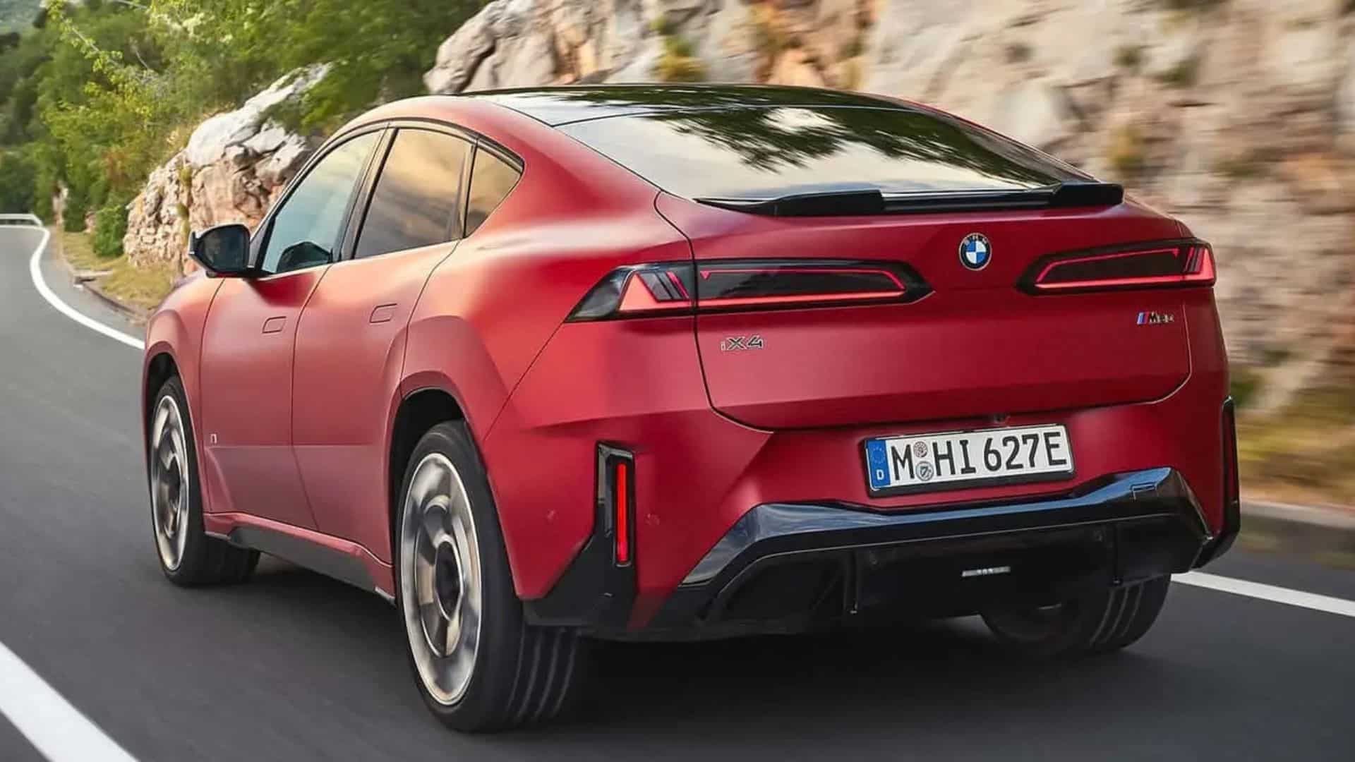 Vista traseira em ângulo do BMW iX4 vermelho em movimento em uma estrada sinuosa de montanha, destacando a silhueta coupé, o spoiler traseiro com luz de freio integrada, as lanternas finas em formato de L e o logotipo BMW no centro da tampa do porta-malas
