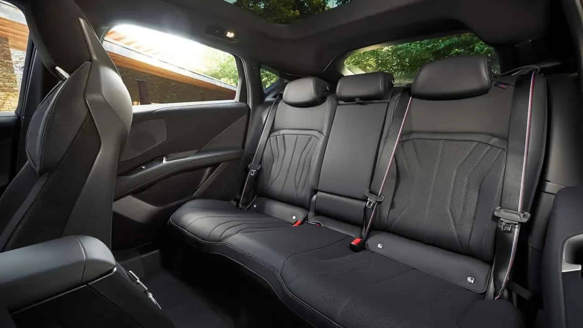 Vista interna do banco traseiro do BMW iX4, com três assentos revestidos em couro preto, detalhes em costura contrastante, apoios de cabeça e cintos de segurança com faixas vermelhas e azuis.