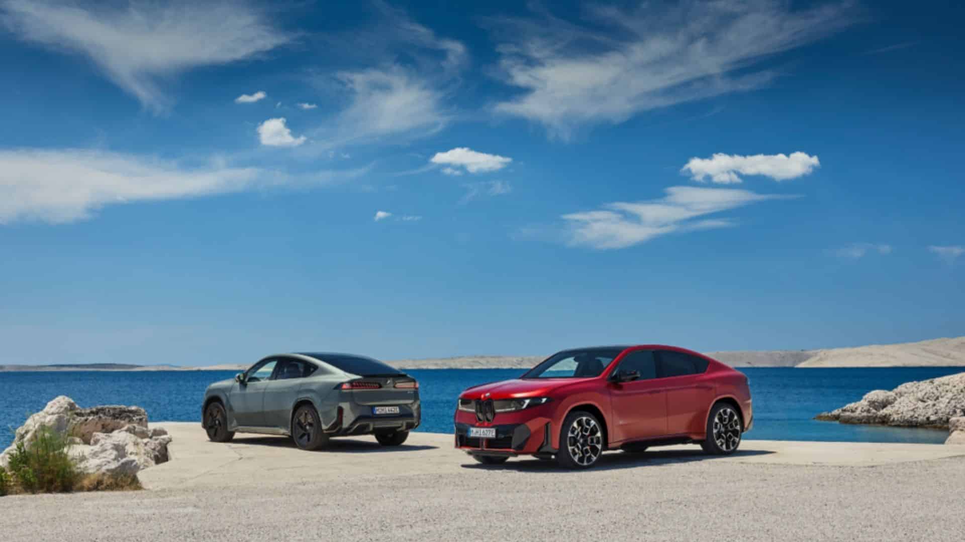 Vista externa de dois BMW iX4 estacionados lado a lado em uma superfície clara à beira-mar, com um modelo vermelho em primeiro plano e outro cinza ao fundo, sob céu azul com nuvens esparsas e montanhas rochosas ao horizonte