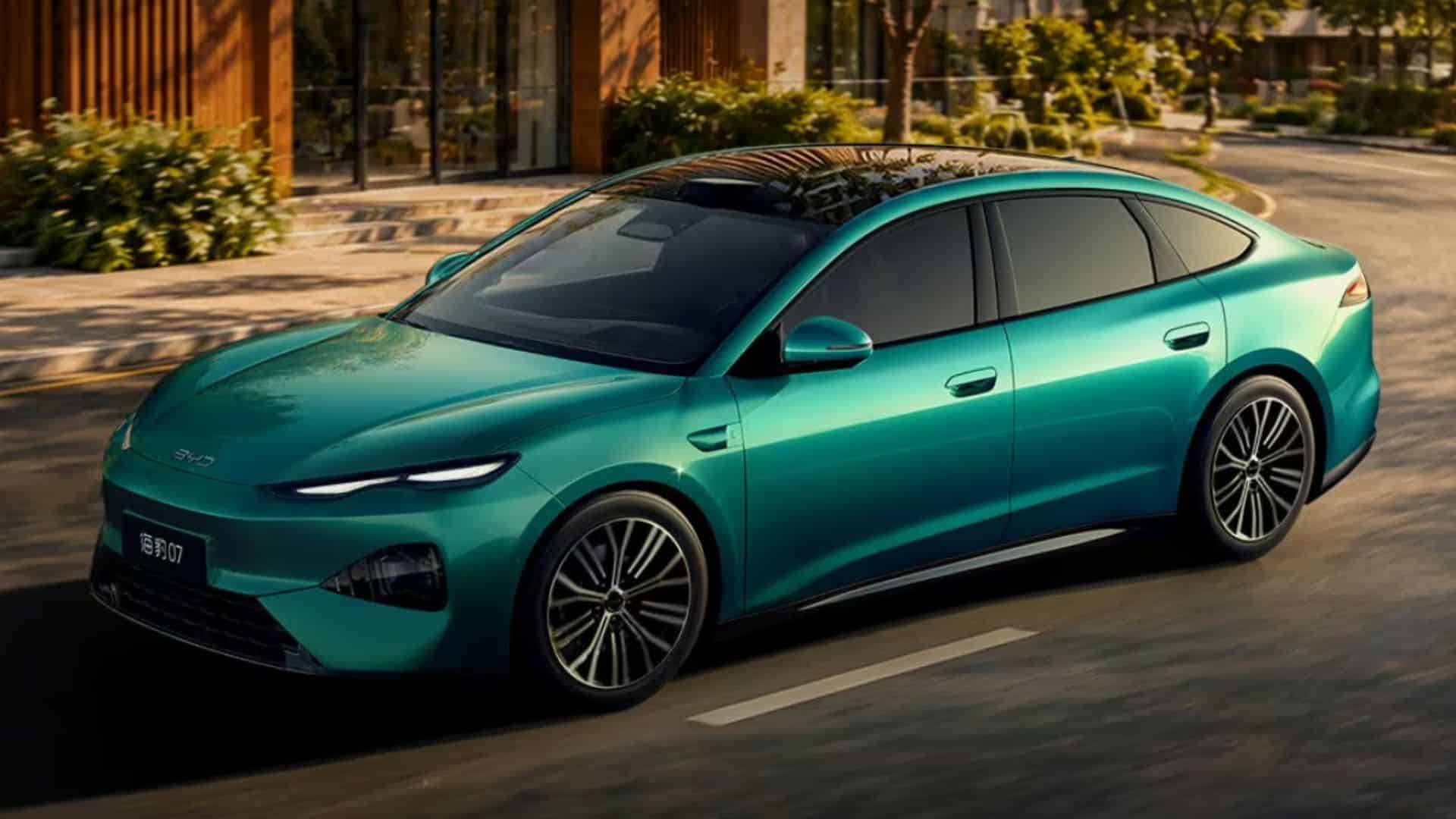 Carro sedã BYD Seal 07 de segunda geração na cor verde, visto em ângulo diagonal frontal. O veículo está em movimento em uma rua asfaltada, com destaque para os faróis divididos em forma de onda, teto panorâmico preto e rodas de liga leve com detalhes em prata