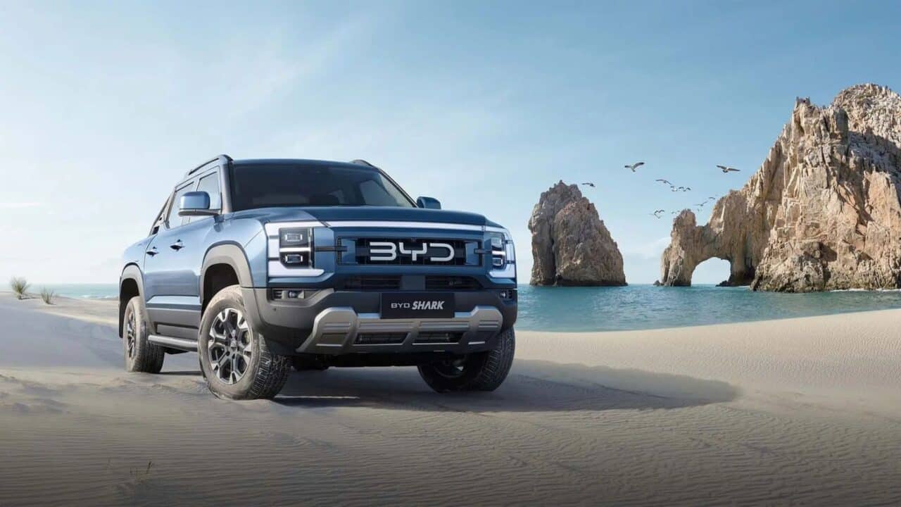 Picape BYD Shark azul estacionada em uma praia de areia clara, com o mar turquesa ao fundo e formações rochosas à direita, incluindo um arco natural