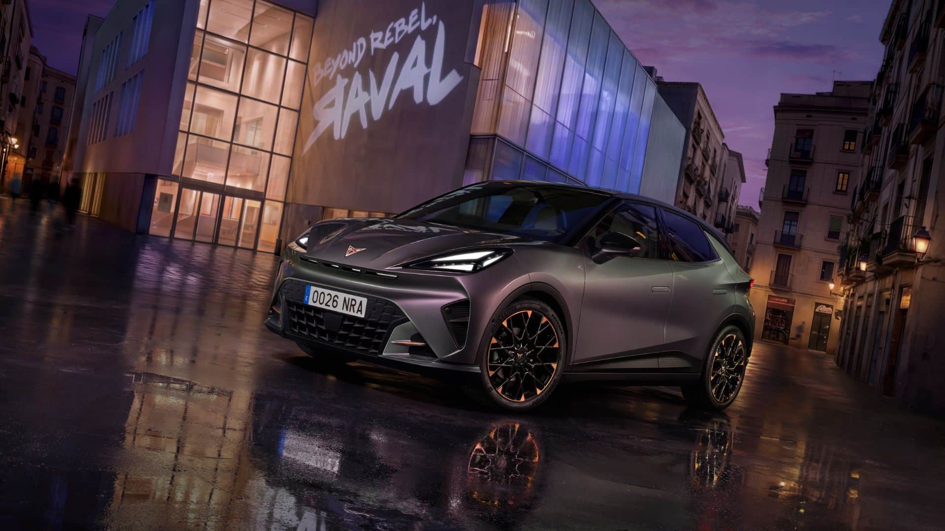 CUPRA Raval cinza-escuro estacionado em uma rua urbana molhada ao anoitecer, refletindo-se no asfalto, com um edifício moderno ao fundo exibindo a inscrição 'BEYOND REBEL RAVAL' em letras grandes