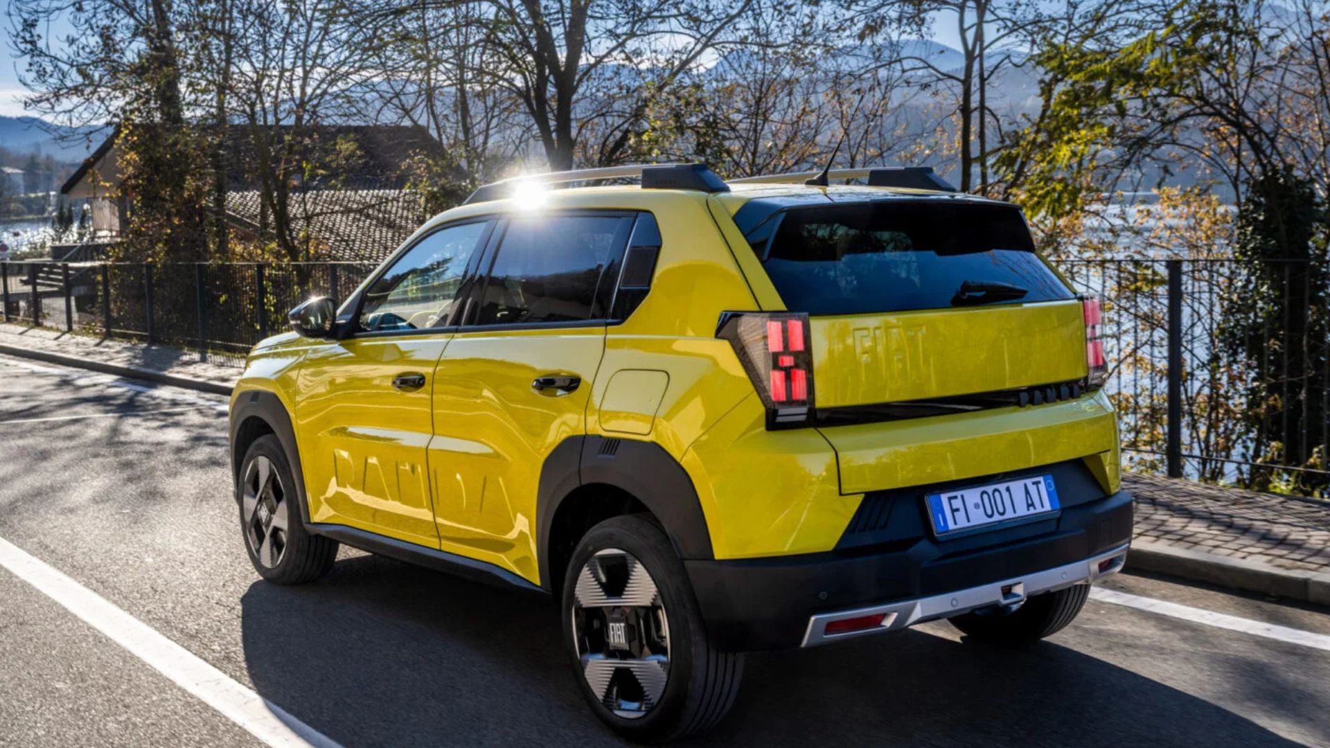 Carro elétrico compacto Fiat na cor amarelo-esverdeado brilhante, fotografado em uma estrada à beira de um lago. A imagem mostra a lateral e a traseira do veículo, destacando a inscrição 'PANDINA' em relevo na porta, detalhes em plástico preto nas caixas de roda, teto com barras longitudinais e lanternas traseiras de LED com formato vertical. A placa italiana azul indica 'FI-001-AT'