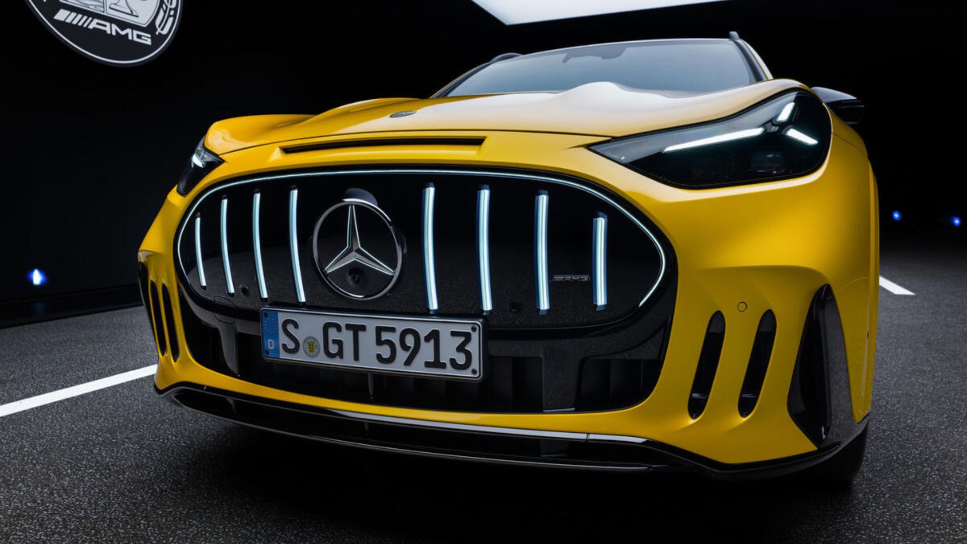 Close-up da dianteira de um SUV esportivo AMG amarelo brilhante em um ambiente escuro, destacando a grade frontal larga com contorno iluminado em LED, o logotipo da Mercedes-Benz ao centro e o emblema AMG à direita, faróis com luzes diurnas afiadas, para-choque agressivo com entradas de ar laterais e a placa de identificação alemã 'S GT 5913