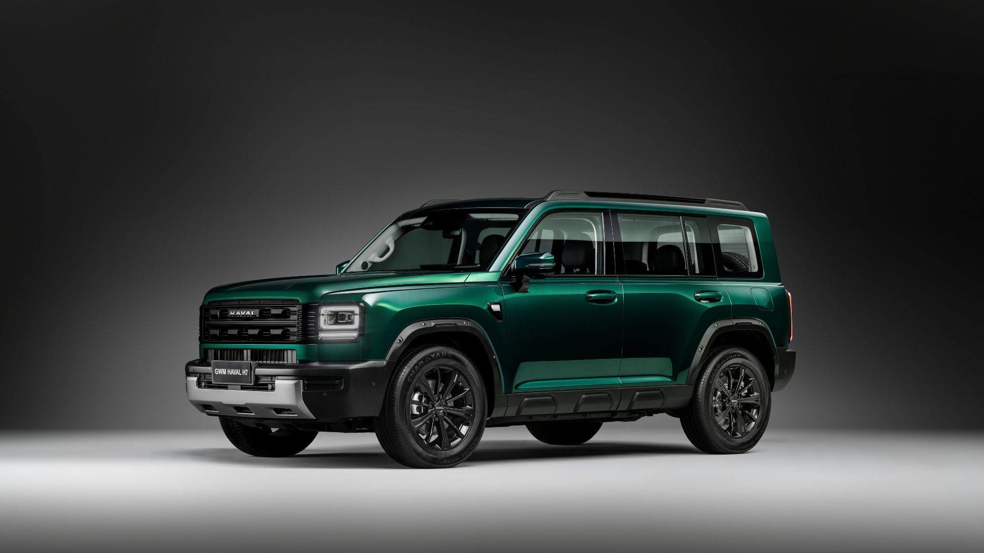 Vista lateral três quartos do GWM Haval H7, mostrando o design quadrado e robusto, teto com barras longitudinais, rodas de liga leve escurecidas, maçanetas convencionais e detalhes em preto nas caixas de roda e saias laterais