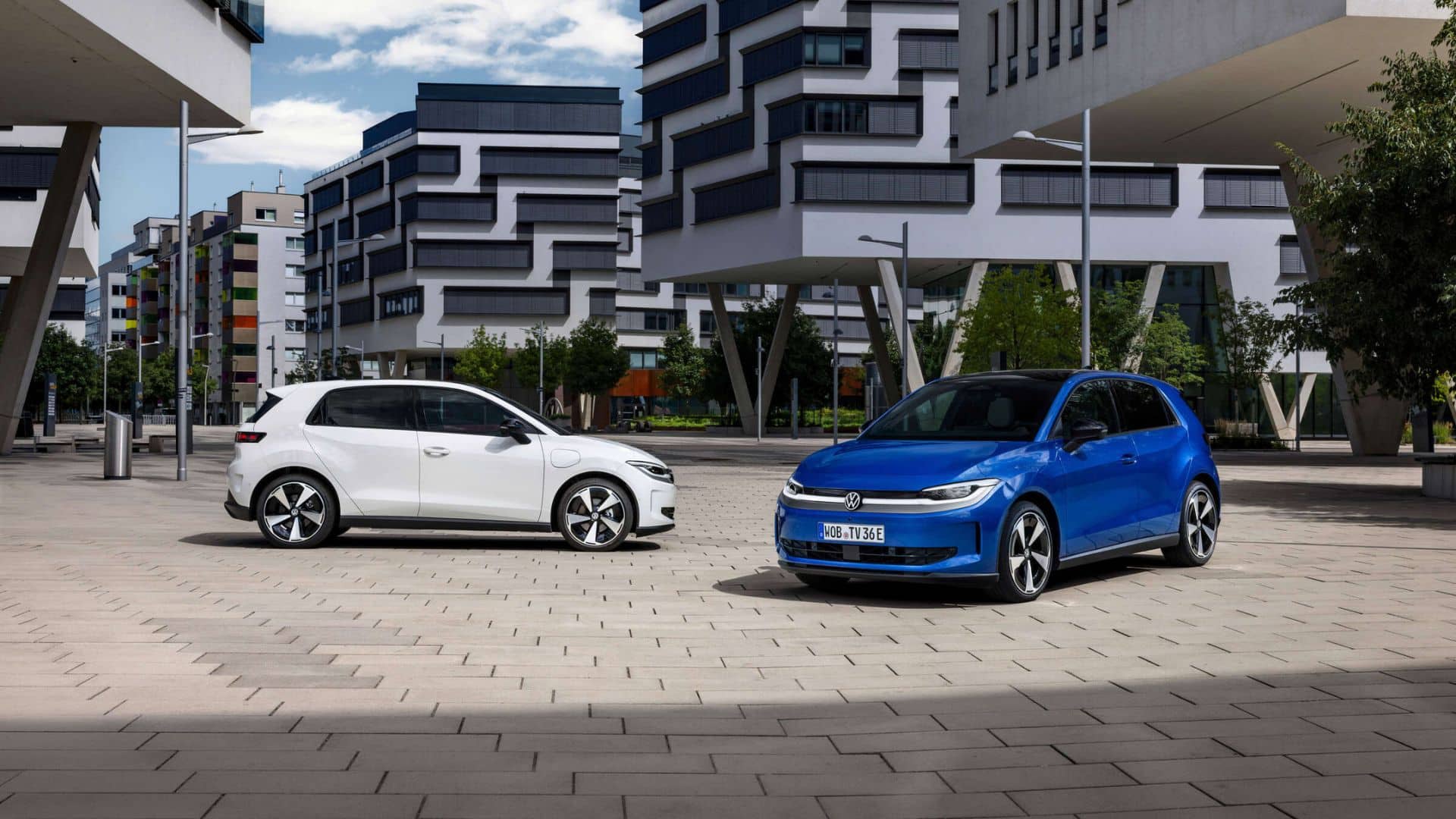 Dois Volkswagen ID. Polo, um branco e um azul, estacionados em uma praça de paralelepípedos claros diante de edifícios modernos de arquitetura geométrica, sob céu azul com nuvens