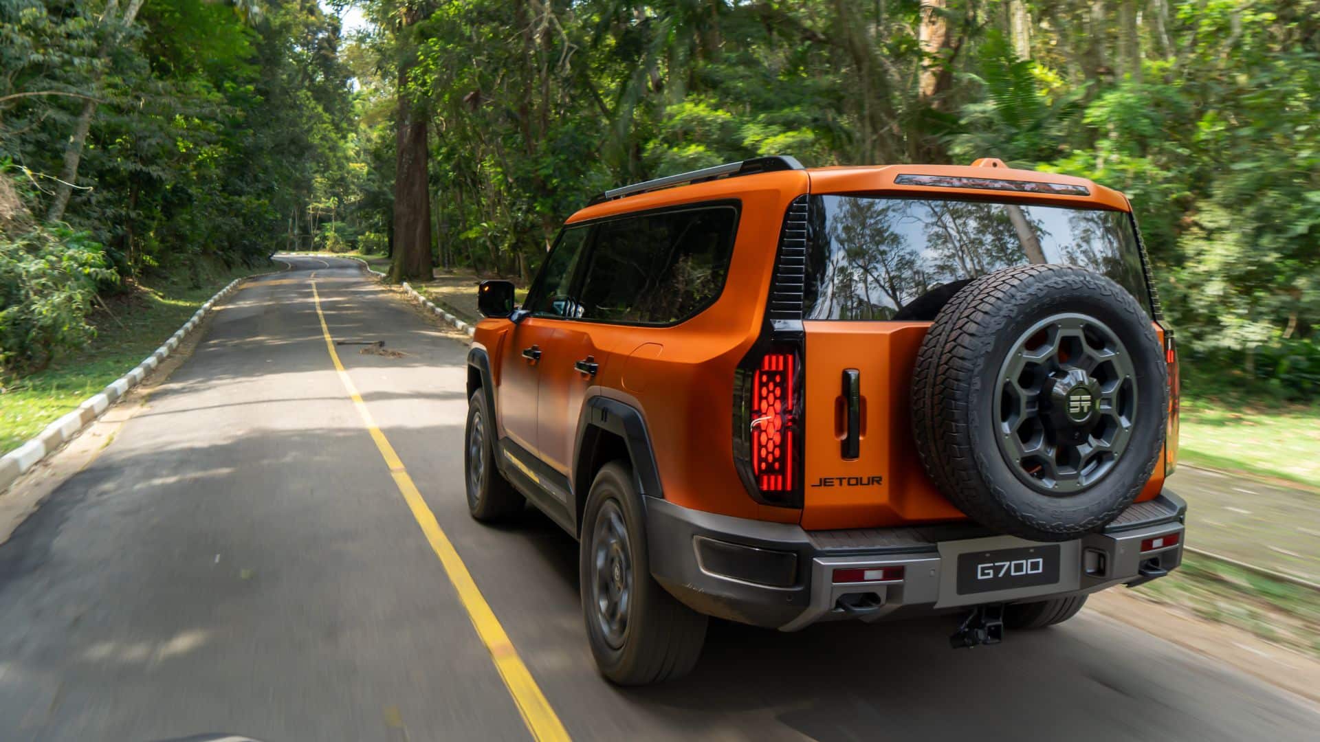 Traseira do JETOUR G700 laranja em movimento em uma estrada asfaltada ladeada por árvores, destacando as lanternas em LED, o estepe na traseira e o nome "JETOUR" na carroceria