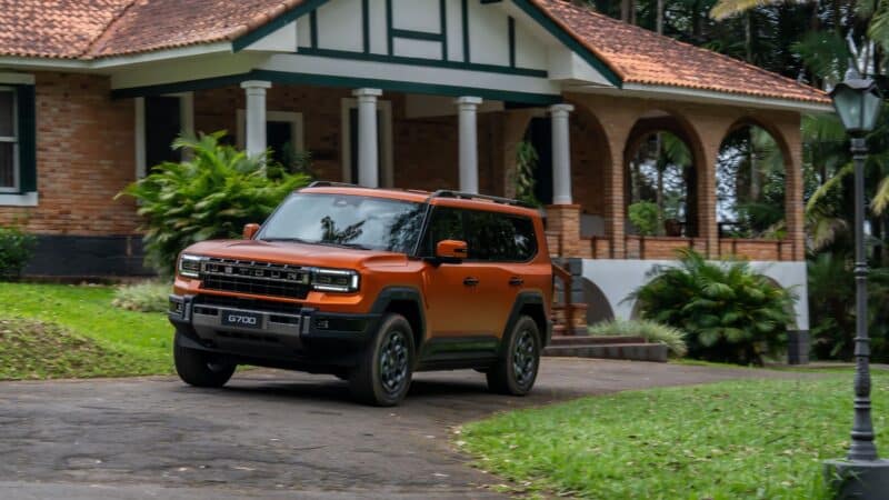 Parte frontal e lateral do JETOUR G700 laranja estacionado em uma entrada de garagem de uma casa de tijolos, com destaque para a grade frontal escura com o nome da marca