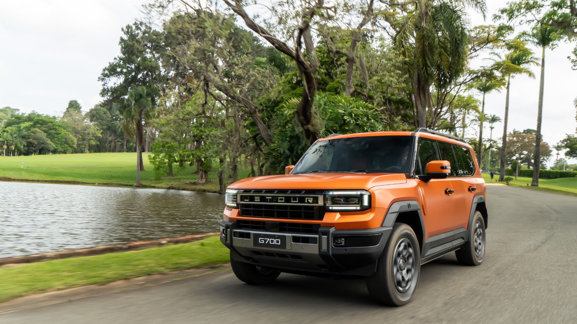 JETOUR G700 laranja em movimento em uma estrada à beira de um lago, com árvores altas ao fundo, destacando o design robusto e a dianteira do veículo