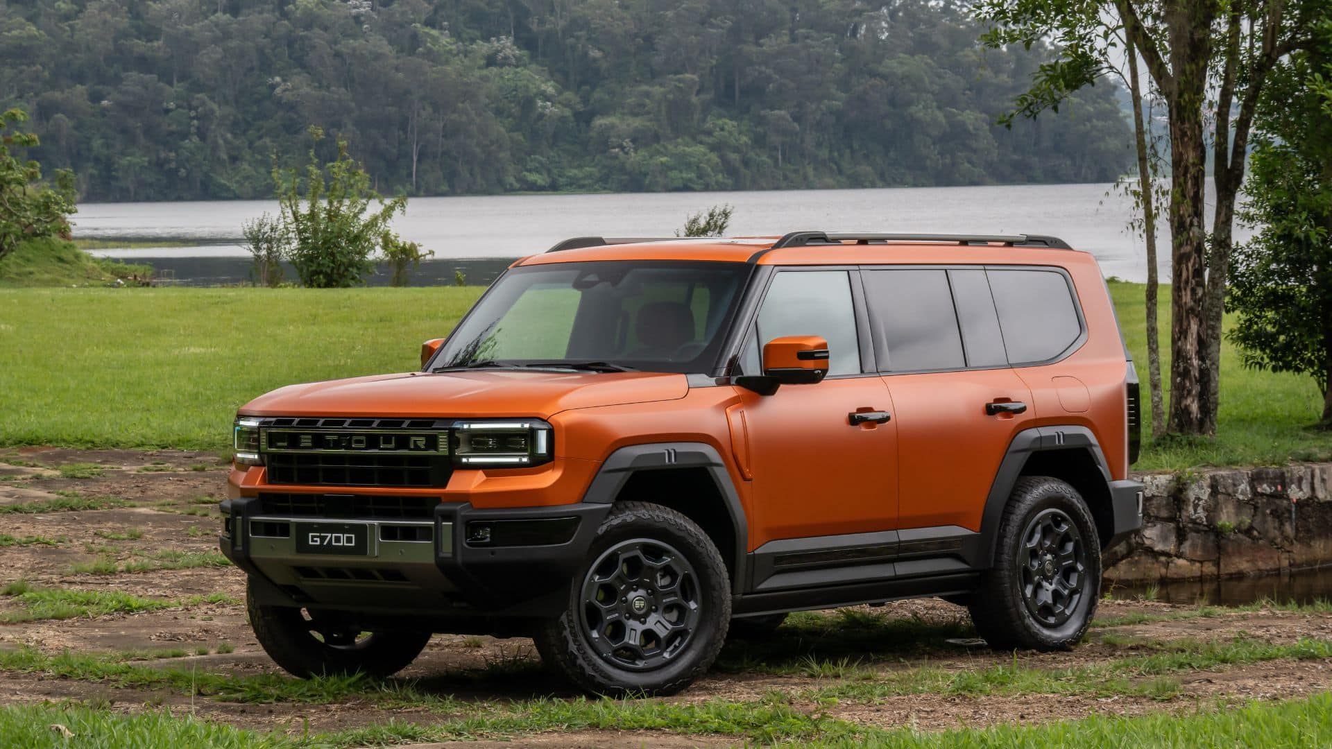 JETOUR G700 laranja em movimento em uma estrada à beira de um lago, com árvores altas ao fundo, destacando o design robusto e a dianteira do veículo
