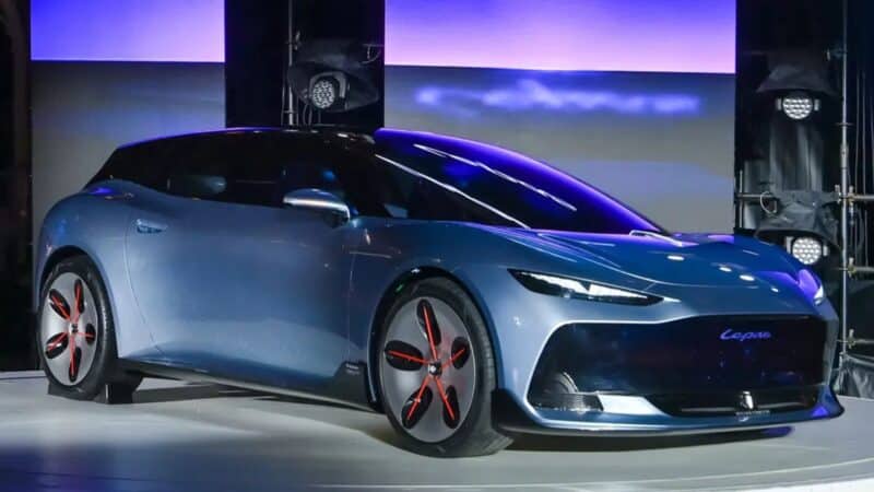 Visão traseira e lateral do LEPAS AURA Concept em um palco. O carro tem carroceria azul clara com teto escuro, lanternas traseiras finas interligadas com o nome 'Lepas' em vermelho, e rodas com detalhes em vermelho. O para-choque traseiro preto exibe a frase 'Drive Your Elegance'