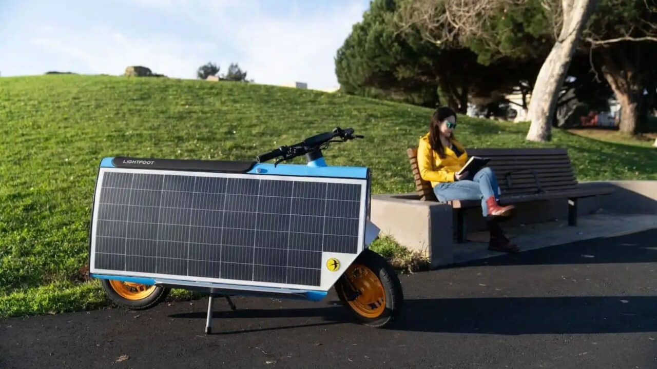 Uma scooter elétrica Lightfoot azul com um grande painel solar retangular preto cobrindo sua lateral, estacionada em uma ciclovia de asfalto ao lado de um gramado verde. Ao fundo, uma mulher de jaqueta amarela sentada em um banco de madeira lê um livro enquanto o sol ilumina a cena