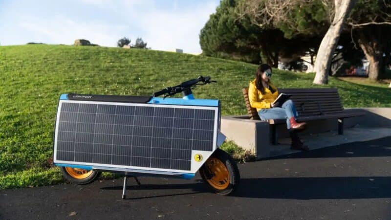 Uma scooter elétrica Lightfoot azul com um grande painel solar retangular preto cobrindo sua lateral, estacionada em uma ciclovia de asfalto ao lado de um gramado verde. Ao fundo, uma mulher de jaqueta amarela sentada em um banco de madeira lê um livro enquanto o sol ilumina a cena