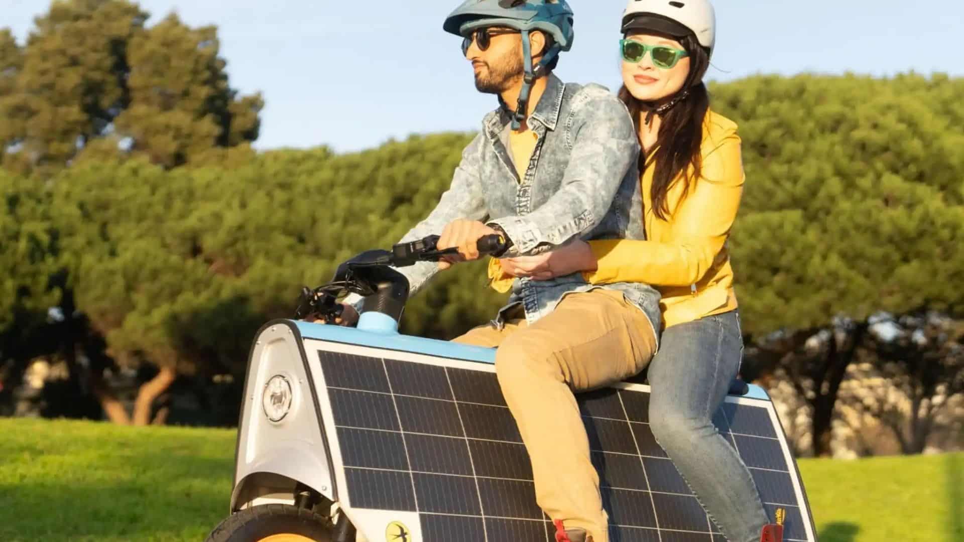 Um homem e uma mulher usando capacetes e óculos escuros andam juntos em uma scooter elétrica Lightfoot com painéis solares nas laterais. O homem, na frente, veste jaqueta jeans e calça caqui, enquanto a mulher, atrás, usa uma jaqueta amarela e abraça a cintura dele. Ao fundo, árvores verdes e um gramado iluminado pelo sol