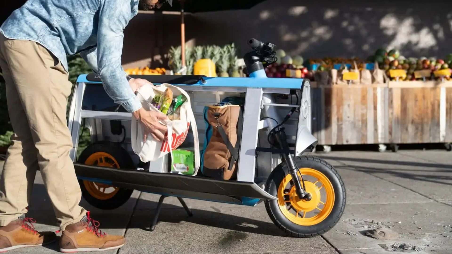 Um homem de jaqueta jeans e calça caqui coloca uma sacola de pano cheia de mantimentos, incluindo vegetais, dentro do compartimento de carga aberto de uma scooter elétrica Lightfoot estacionada. A scooter tem painéis solares nas laterais e rodas com aro amarelo, e ao fundo há uma barraca de feira com frutas e legumes expostos