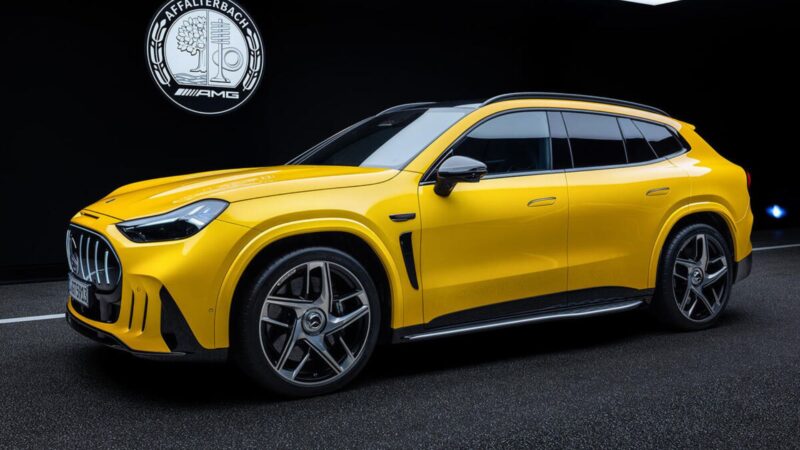 Vista lateral e dianteira de um SUV esportivo amarelo brilhante com grade frontal vertical iluminada, faróis afiados, detalhes aerodinâmicos escuros nas laterais e rodas de liga leve de grande porte, posicionado sob um logotipo histórico da AMG ao fundo
