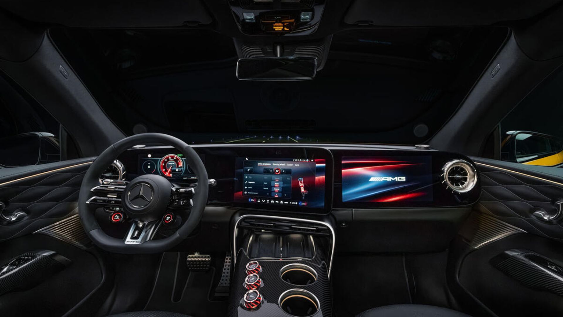 Painel interno de um carro esportivo de luxo com volante AMG, três telas digitais integradas (instrumentos, central multimídia e tela do passageiro exibindo o logo AMG), saídas de ar redondas e console central em fibra de carbono com botões vermelhos de controle