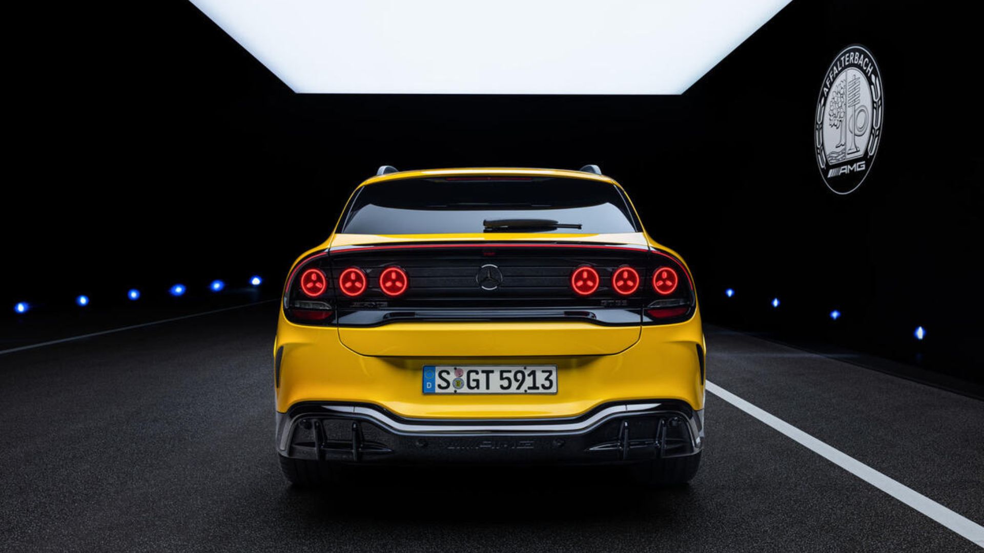 Vista frontal traseira de um SUV AMG amarelo em um ambiente escuro, exibindo a barra de luz traseira contínua, o logotipo da Mercedes ao centro, o para-choque agressivo com difusor de fibra de carbono e a placa de identificação 'S GT 5913'