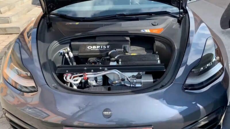 Vista frontal de um Tesla Model 3 cinza com o capô aberto, revelando um motor a gasolina da Obrist Powertrain instalado no compartimento dianteiro (frunk), com componentes mecânicos, mangueiras e a tampa preta do motor com a inscrição 'OBRIST POWER TRAIN
