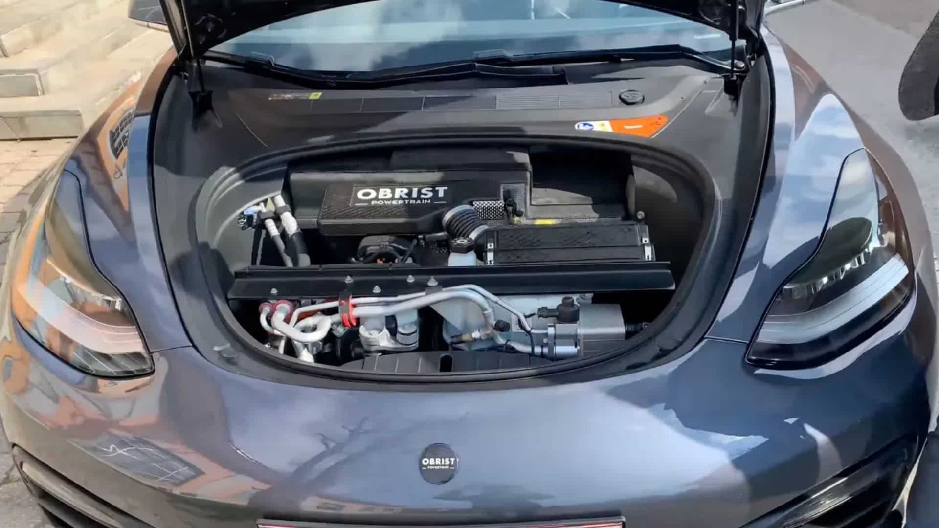 Vista frontal de um Tesla Model 3 cinza com o capô aberto, revelando um motor a gasolina da Obrist Powertrain instalado no compartimento dianteiro (frunk), com componentes mecânicos, mangueiras e a tampa preta do motor com a inscrição 'OBRIST POWER TRAIN