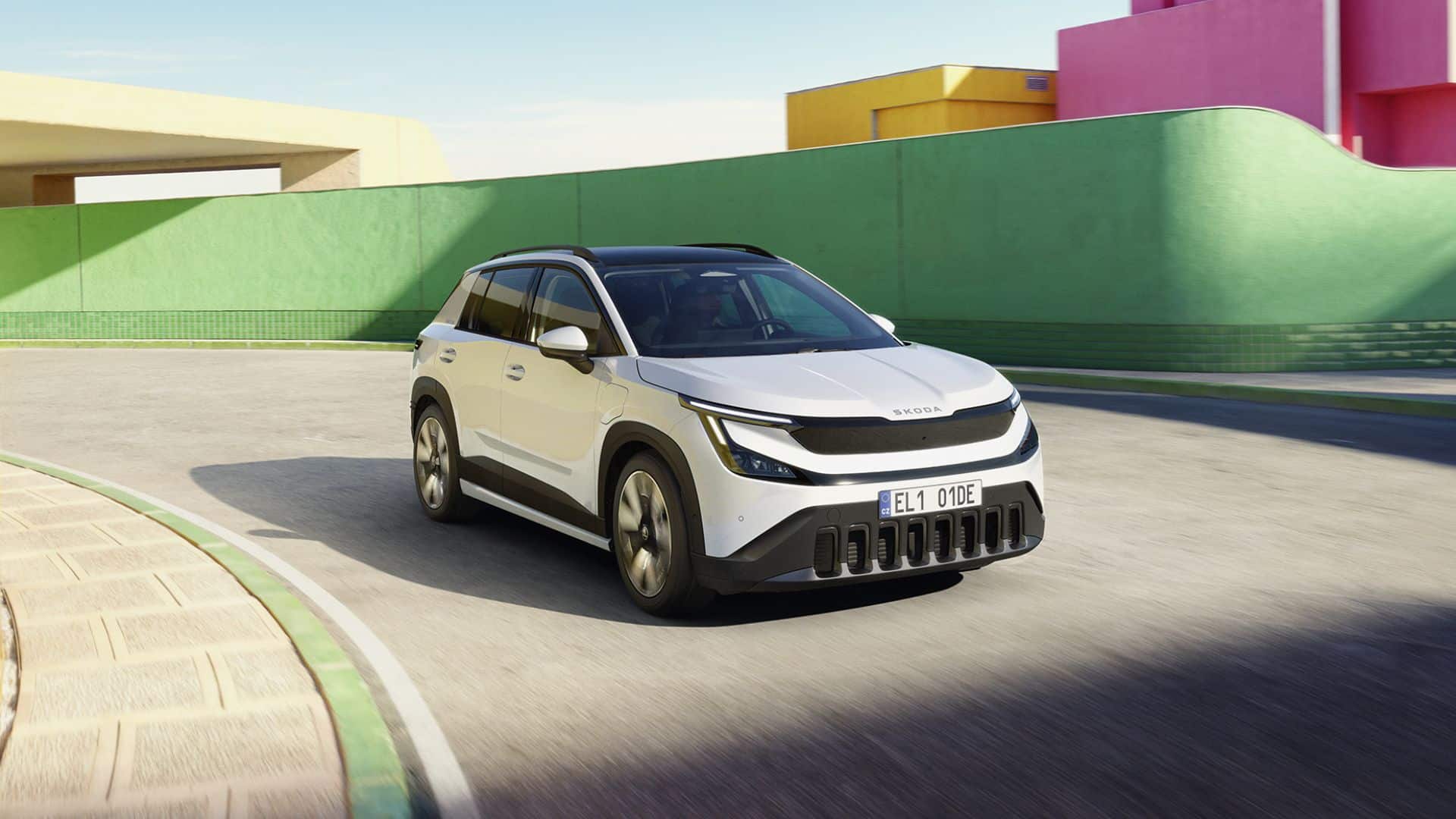 Skoda Epiq branco com teto preto em movimento em uma curva de rua urbana, com muros coloridos em verde, amarelo e rosa ao fundo, destacando a frente com grade escura, faróis afilados e o nome 'SKODA' no capô