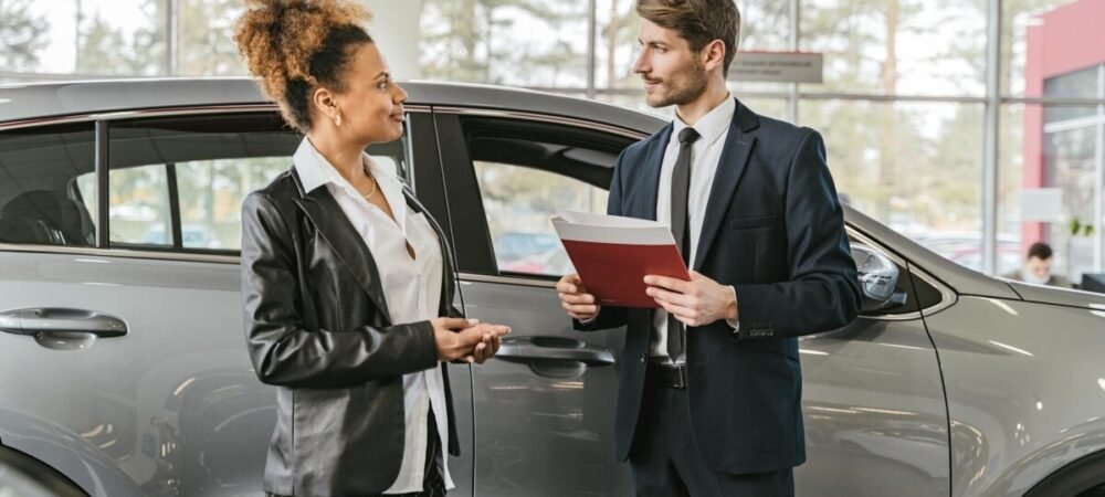 Carro elétrico sendo negociado na concessionária, uma mulher conversando com um homem segurando uma pasta em frente a um carro na concessionária
