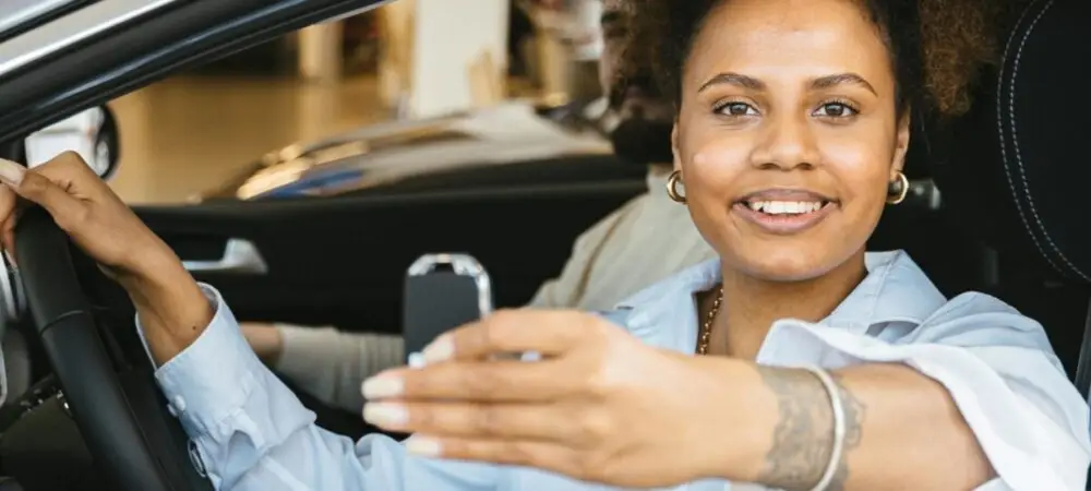 Imagem mostrando mulher num carro dentro de uma concessionária, segurando a chave na mão, sorrindo, indicando que comprou o carro